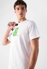 Balta medvilnė marškinėliai, turintys didelį žalią ampersand'ą ir prekės ženklą krūtinėje. Modelis laiko pilką diržą per petį.