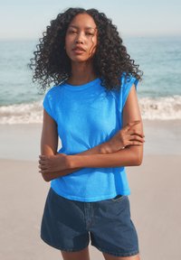 Bright blue short-sleeve t-shirt with a round neckline, paired with dark denim shorts, against a beach background with waves.