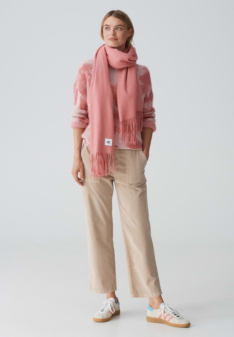 Roze sjaal met franjes, gedragen over een licht gebreide trui met patroon, gecombineerd met beige wijde broekspijpen en witte sneakers met gekleurde accenten.