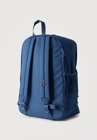 Μπλε σακίδιο πλάτης Jansport με επενδυμένους ιμάντες ώμου, θηλιά για κρατήρα και πλαϊνή τσέπη από δίχτυ, σε απλό φόντο.