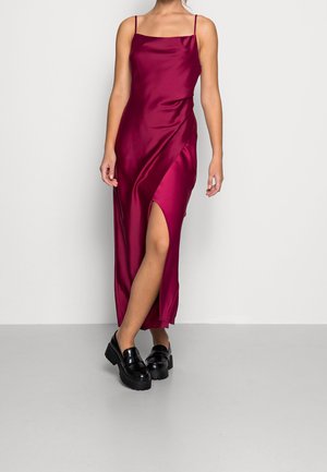 Vrouw die een mouwloze bordeauxrode satijnen jurk draagt met een hoge split aan de dij, gecombineerd met zwarte chunky loafers, staand voor een effen achtergrond.