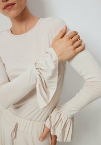 Žena v svetložltom topánke s dlhými rukávmi a plisovanými volánovými manžetami, s rukou položenou na opačnom ramene, v kombinácii so zodpovedajúcimi béžovými nohavicami.