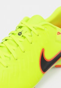 Neon gul fodboldsko med tekstureret syntetisk overdel, sort og orange Nike swoosh samt matchende snørebånd. Nærbillede fremhæver design detaljer.