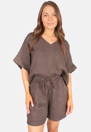 Woman with long brown hair wearing a loose brown V-neck top and matching drawstring shorts standing against a plain background.