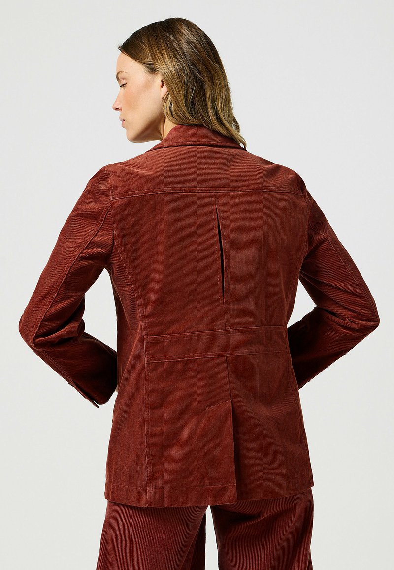 Vrouw met lichtbruin haar, gekleed in een roestkleurige corduroy blazer en bijpassende broek, van achteren weergegeven tegen een effen achtergrond.