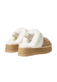 Een paar tan gekleurde suède platformslippers met dikke witte fleece voering en een decoratieve band over de achterkant van de hak.