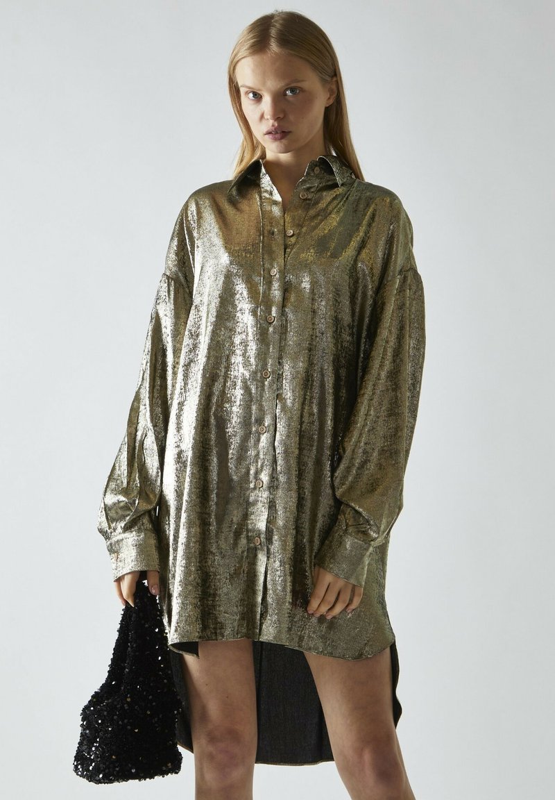 Glänzendes, metallisch goldfarbenes, übergroßes Hemd mit langen Ärmeln, durchgehender Knopfleiste, spitzem Kragen und geschwungenem Saum, kombiniert mit einer schwarzen Pailletten-Tasche.