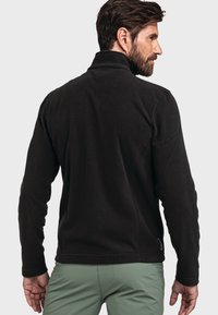 Hombre con barba que lleva una chaqueta de forro polar negra visto desde atrás, combinado con pantalones verdes claros, contra un fondo liso claro.