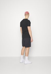 Black short-sleeve t-shirt with a fitted design, paired with dark shorts. Man stands in white sneakers and knee-high socks, viewed from behind.