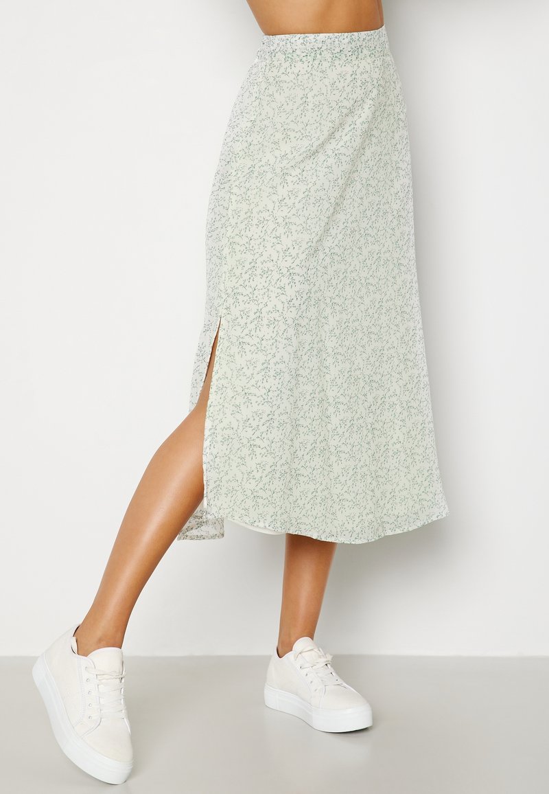 Falda midi verde claro con estampado floral y una abertura lateral, hecha de una tela ligera y fluida. Combinada con zapatillas blancas.