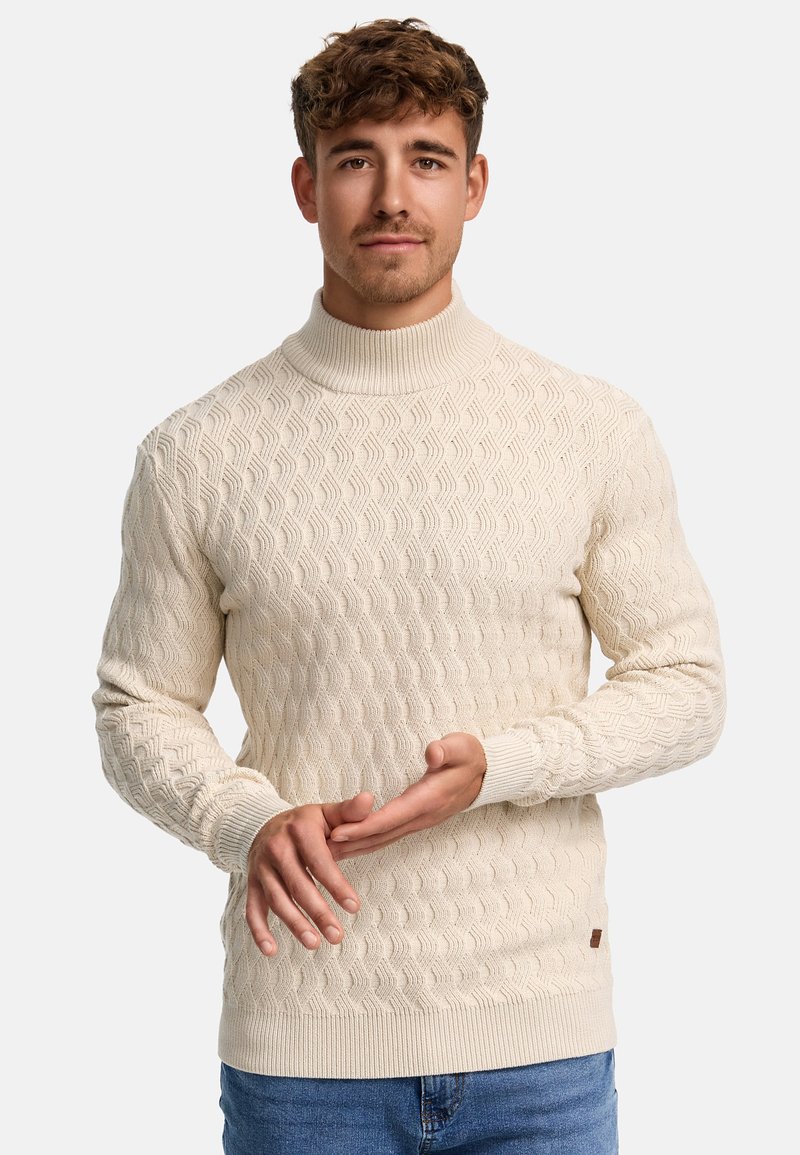 Pull en tricot couleur crème avec un col montant, arborant un motif texturé en vagues et des poignets côtelés. Détail subtil de l'étiquette de la marque à l'ourlet.
