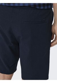 Marineblå bomullsshorts med en glatt tekstur, en baklomme og en ren finish i kanten. Synlig sømdetalj på siden.