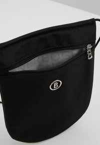 Svart tygväska med en framfickas blixtlås, silverfärgade detaljer och präglad logotyp. Slät yta med en minimalistisk design.