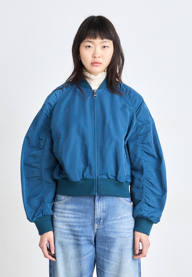 Chaqueta bomber en color verde azulado con cierre frontal, puños y dobladillo acanalados, y mangas fruncidas. Combinada con jeans de color azul claro, modelo mirando al frente.