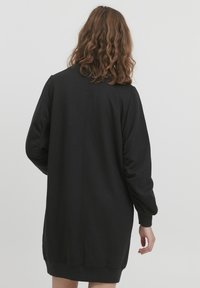 Abito felpa nero oversize con maniche lunghe, polsini e fondo a costine, caratterizzato da una cucitura centrale sul retro e da un tessuto morbido e testurizzato.