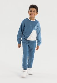 Kék pamut pulóver fehér betéttel; hozzáillő kék melegítőnadrág. Mindkettő bordázott mandzsettával ellátott. A modellen fehér sportcipő van.