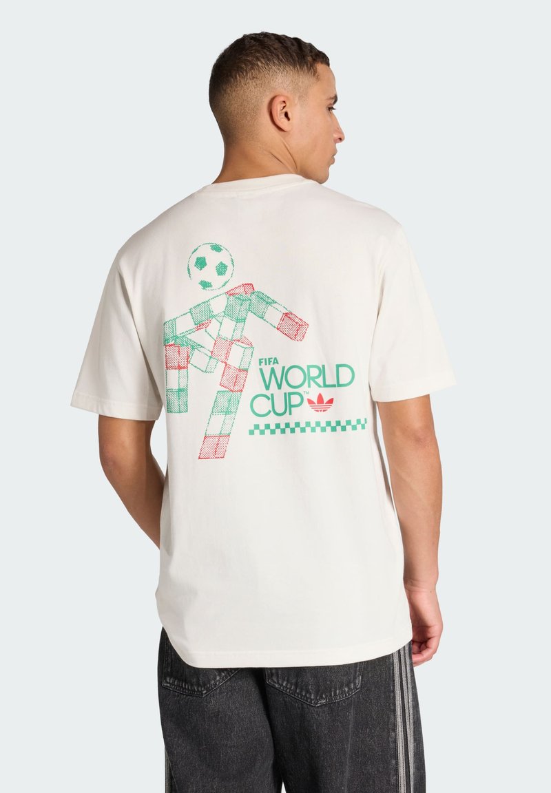 Uomo con maglietta bianca con giocatore di calcio stilizzato pixelato e scritta "FIFA World Cup" sul retro, abbinata a jeans scuri.