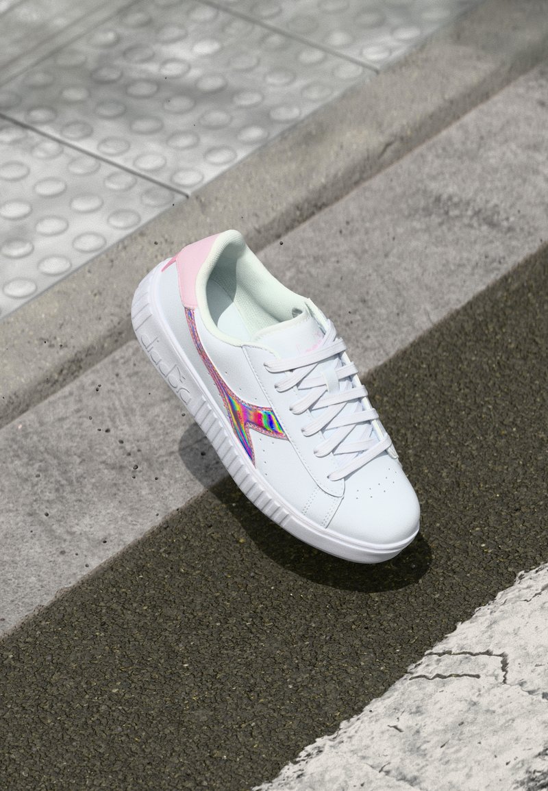 Fehér sneaker rózsaszín kiegészítőkkel, sima szintetikus anyagból, klasszikus fűzős kialakítással és szivárványos oldalsó részletekkel. Strukturált talp.