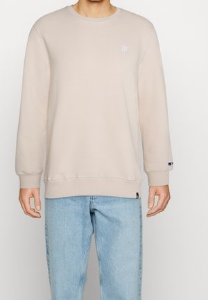 Homme portant un sweat-shirt beige à col rond et un jean bleu clair, debout, les bras détendus le long du corps, sur un fond uni.