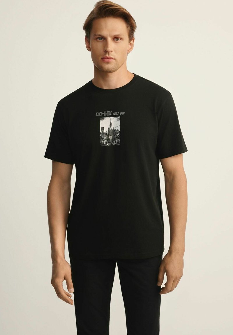 Czarna bawełniana koszulka z grafiką przedstawiającą panoramę miasta z napisem "OCHNIK EST. 1999" powyżej. Krótkie rękawy, okrągły dekolt.