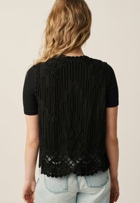 Vesta brodată neagră are un material texturat cu linii verticale și accente florale, cu o margine detaliată și pompoane, purtată peste un top cu mânecă scurtă.