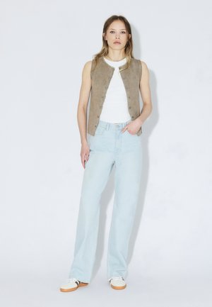 Beigenvärinen hihaton liivi valkoisen topin päällä, yhdistetty vaaleansinisiin leveälahkeisiin farkkuihin ja valkoisiin lenkkareihin, joissa on vaaleanruskea pohja; yksinkertainen, selkeä muotoilu.