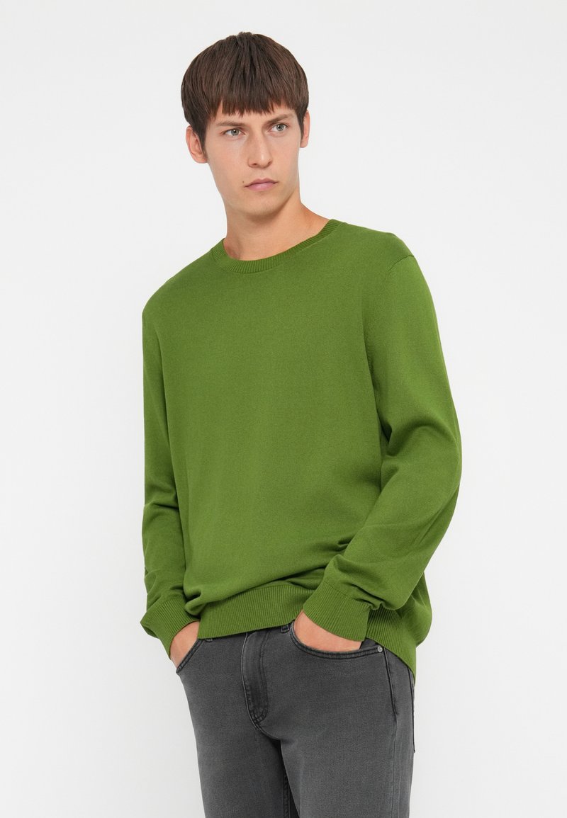 Zielony wełniany sweter z okrągłym dekoltem i ściągaczami. W połączeniu z ciemnoszarymi spodniami, model stoi z rękami w kieszeniach na jednolitym tle.