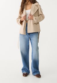 Veste beige légère avec col et détail de nœud, associée à un jean bleu clair à jambes larges avec un dégradé progressif. Baskets marron.