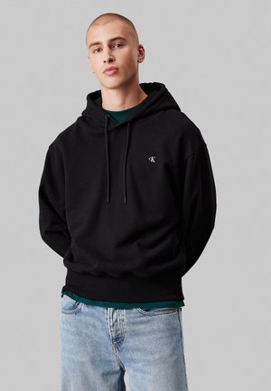 Felpa nera in cotone con cappuccio regolabile e un piccolo logo sul petto. Abbinata a jeans azzurri chiari, con una texture liscia.