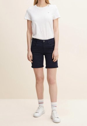 Person iført en hvid kortærmet t-shirt, mørke opkrogede shorts, hvide sneakers og hvide sokker med røde og blå striber.