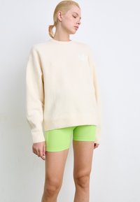 Crèmekleurige oversized trui met geribbelde manchetten, gecombineerd met strakke neongroene bike shorts. Soepele texturen, minimale logodetail.
