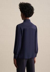 Blusa navy con colletto alto, maniche a sbuffo e polsini con bottoni. Tessuto liscio, vestibilità ampia e design pulito sulla parte posteriore.
