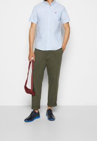Camicia azzurra a maniche corte con logo rosso, pantaloni verde oliva con risvolto, borsa regolabile bordeaux e scarpe nere con suola blu.