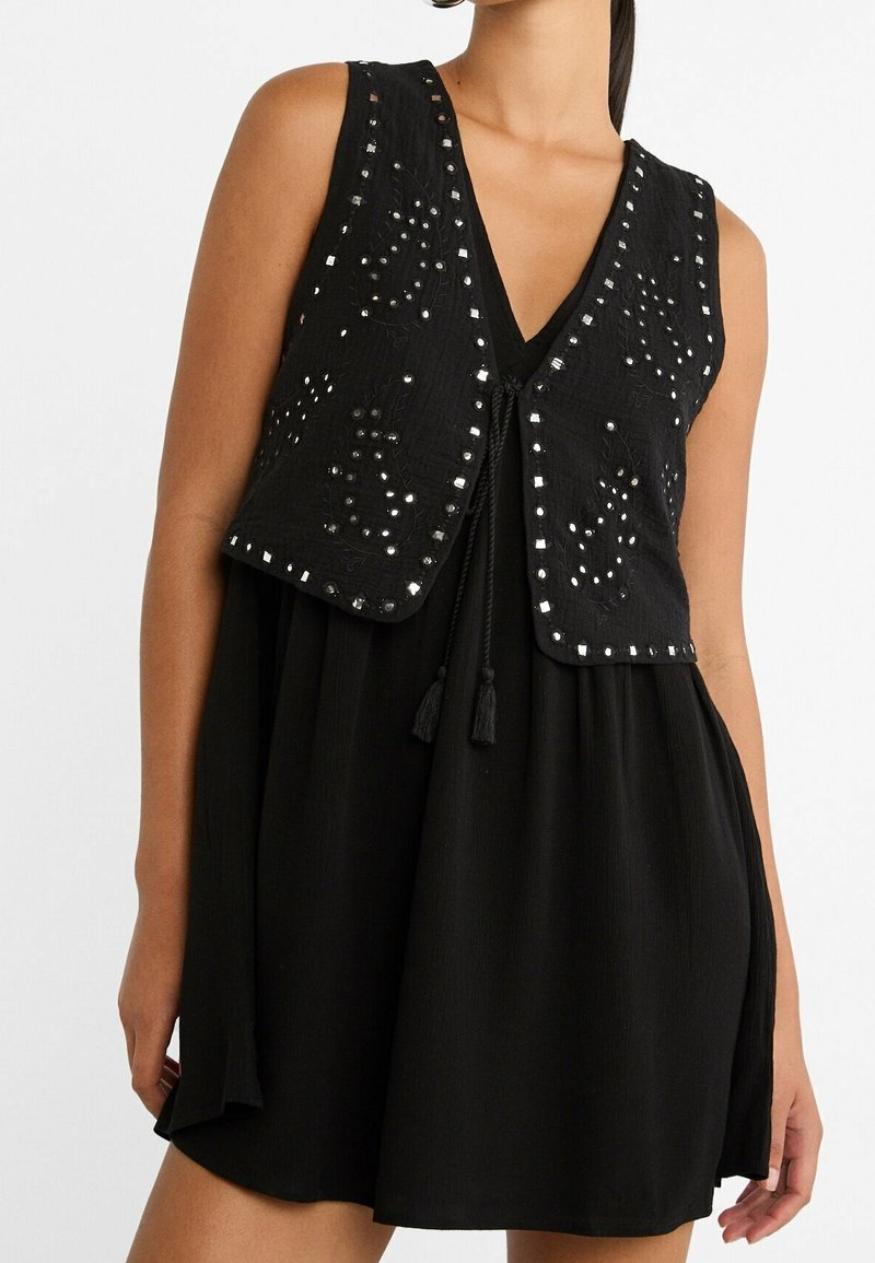 Femme portant une robe noire sans manches avec un gilet noir sans manches orné de clous réfléchissants et de broderies.