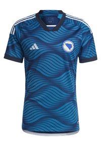 Niebieska koszulka piłkarska z falistymi wzorami, krótkimi rękawami, dekoltem w serek, trzema białymi paskami na ramionach oraz okrągłym logo drużyny na piersi.