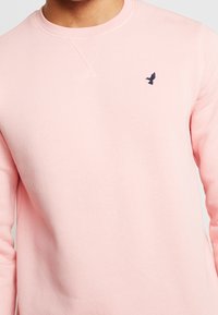 Felpa leggera rosa con scollo rotondo, maniche lunghe e un piccolo logo di un uccello blu navy sul lato sinistro del petto. Tessuto morbido, vestibilità standard.