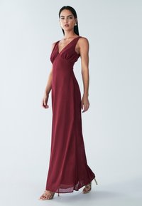 Vínové maxi šaty s výstrihom do V, zdobené nariaseným detailom na hrudi a ľahkým splývavým strihom. Majú nastaviteľné ramienka a bočný rozparok.