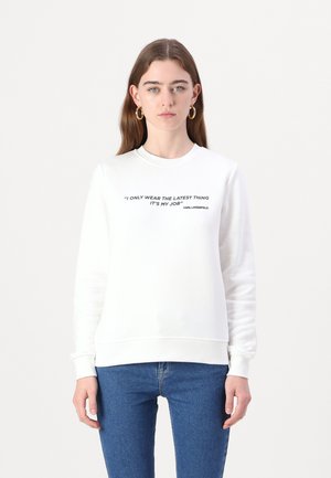 Hvit genser med svart trykt sitat sentrert på brystet, rund halslinning og tilpassede mansjetter, kombinert med blå jeans.