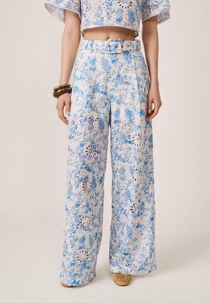 Mujer que lleva pantalones de talle alto y pierna ancha y un top corto a juego con estampado floral azul y detalles con ojales, zapatos beige y pulseras en la muñeca derecha.