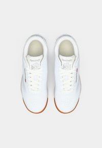 Par de sapatilhas brancas Reebok Classic com solas de borracha e atacadores brancos, vistas de cima sobre um fundo branco.