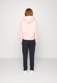 Ljusrosa hoodie med huva och ribbade ärmslut, ihop med navy-randiga byxor och vita sneakers, sedd bakifrån.