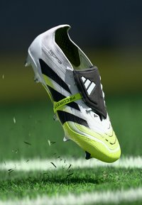 Bijela i neonska žuta Adidas Predator nogometna kopačka u zraku iznad travnatog terena s crnim trakama i zaštitnim poklopcem na vezicama.