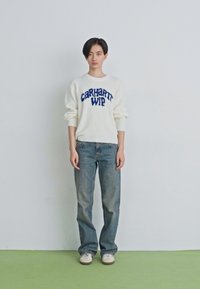 Inimene seisab valge seina ääres, seljas valge Carhartt WIP sviiter, sinised teksad ja valged tennisetossud rohelisel põrandal.