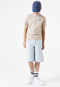T-shirt en coton beige avec du texte, short en jean bleu clair, baskets blanches et chaussettes à motifs noir et blanc. Casquette bleue sur la tête.