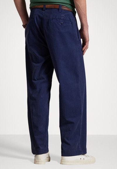 Pantalons en coton bleu marine avec une coupe décontractée, deux poches arrière et une ceinture en cuir marron visible. Les jambes se resserrent légèrement au niveau de la cheville.