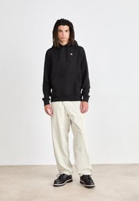Sudadera negra con bolsillo frontal y cordón, combinada con pantalones beige claros. Zapatos negros con detalle contrastante en la suela. Diseño simple.