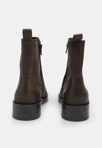 Botas de cuero marrón hasta el tobillo con una textura suave, con cremalleras laterales y un tacón bajo de goma, vistas desde un ángulo trasero.
