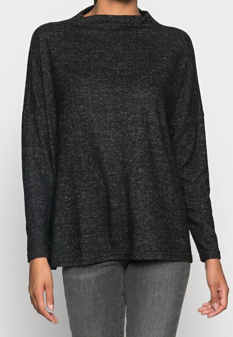 Femme portant un pull ample à manches longues gris foncé et un jean gris clair, debout devant un fond clair uni.
