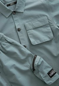 Chemise bleu clair à boutons avec une poche poitrine unique, poignet élastique et poche latérale zippée, fabriquée en tissu lisse.