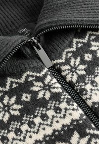 Grijs gebreid vest met een ritsluiting, voorzien van een zacht geribbeld kraag en een wit sneeuwvlokpatroon op een donkere achtergrond.